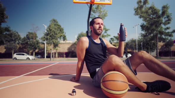 Tired Basketball Player After the Match Sits on the Basketball Field and Drinks Water