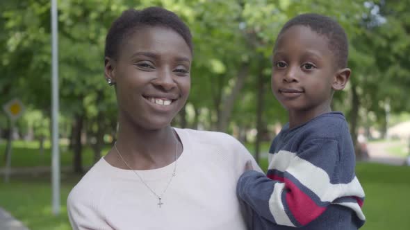 Portrait of Adorable African American Woman Holding Her Funny Son in Her Arms in the Green Park