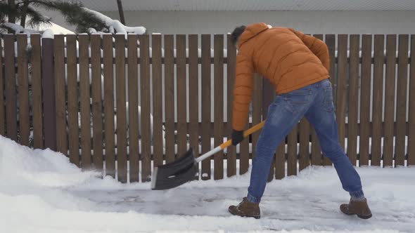A man shovels snow from the road near the fence with a snow shovel.