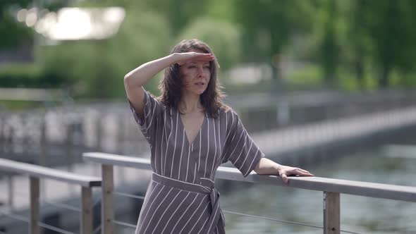 Portrait of a Brunette in a Striped Dress at the Railing of a Bridge Over the River on a Windy Sunny