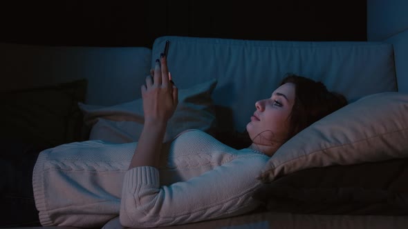 Millennial Woman Laying In Bed Holding Cellphone, Swiping Photos In Online Dating App 