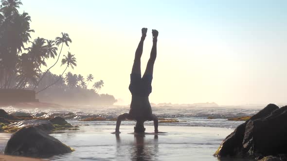 Athlete Stands on Hands and Head Against Foaming Ocean Waves