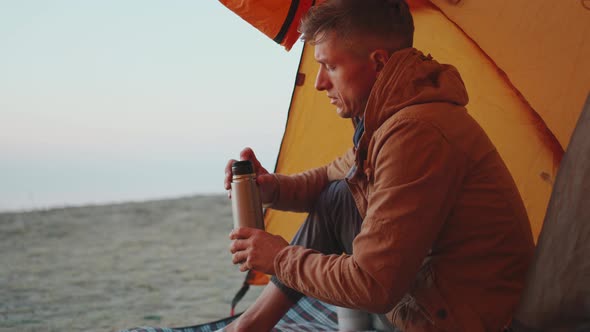 Handsome Adult Man Hiker Sitting in Tent in Casual Wear and Pouring Warm Coffee From Thermos in Mug