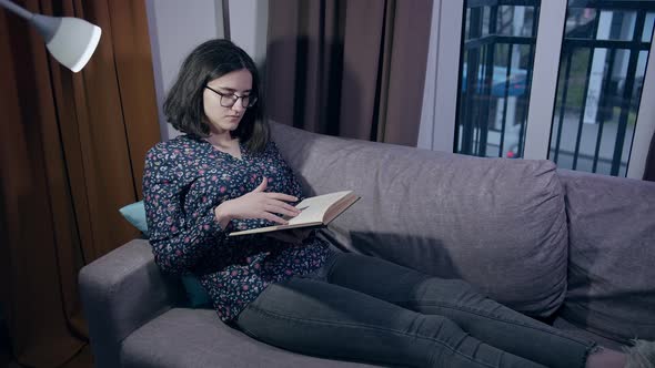 Girl in Glasses Sits on the Couch in the Evening with a Book Cars Drive Outside the Window