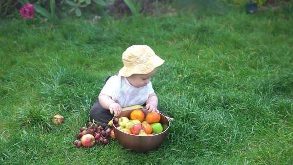 Small Newborn Child in Summer Panama Hat Sit on Grass Barefoot in Bib with Big Bowl of Fresh Fruit