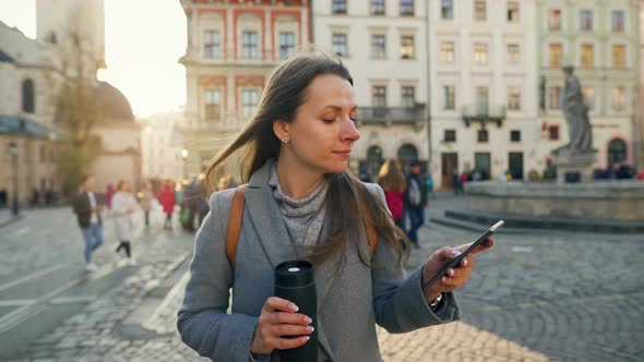 Woman with a Thermos Cup in Hand Walking Down an Old Street Using Smartphone at Sunset