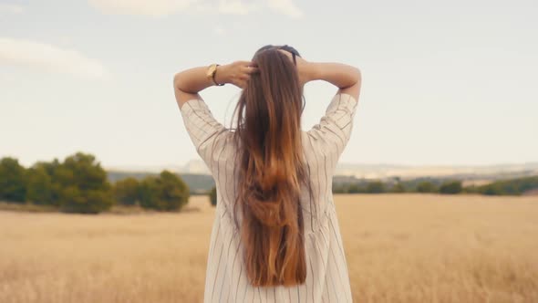 Brunette Woman Tying Her Long Blonde Hair While Standing In The Meadow - Back View -