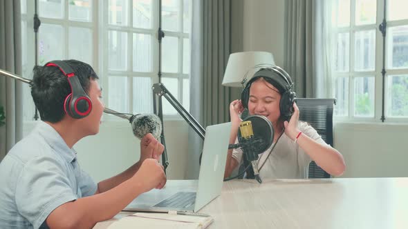 Asian Kid Boy In Headphones Singing Into Microphone While Recording Podcast With Boy Host In Studio