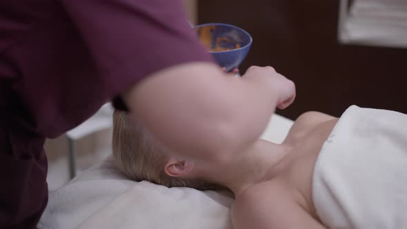 Beautiful Caucasian Woman with Closed Eyes Lying in Spa Salon As Cosmetician Applying Honey Mask on