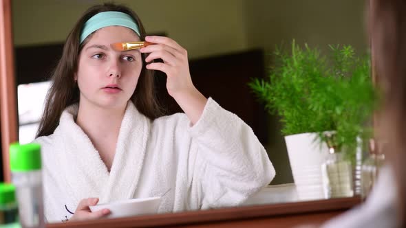 miling young woman applying clay facial mask looking in bathroom mirror