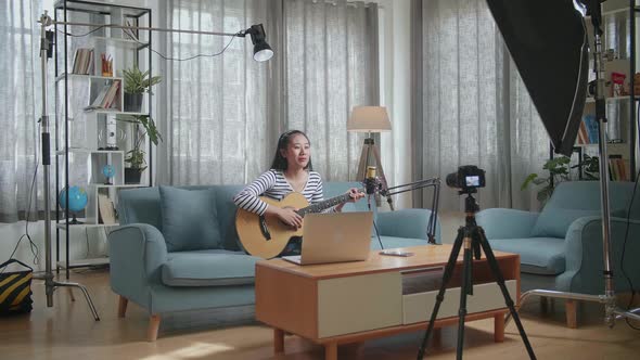 Asian Woman With A Laptop Waving Hand And Speaking To Camera While Live Stream Playing A Guitar