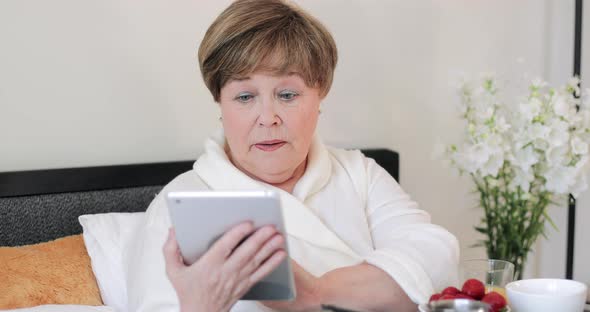 Old Lady with Serious Face Expression Wearing Home Rob and Using Tablet. Elderly Female Pensioner