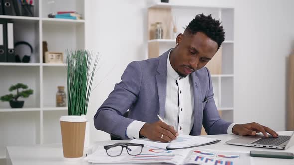 Young Male Office Worker Typing on Laptop and Writing Down to His Report Important marks