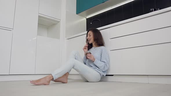 Smiling Woman in Stylish Clothes Sitting on the Kitchen Floor and Eating Chocolate Cream From Jar