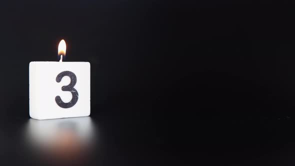 A square candle saying the number 3 being lit and blown out on a dark black background