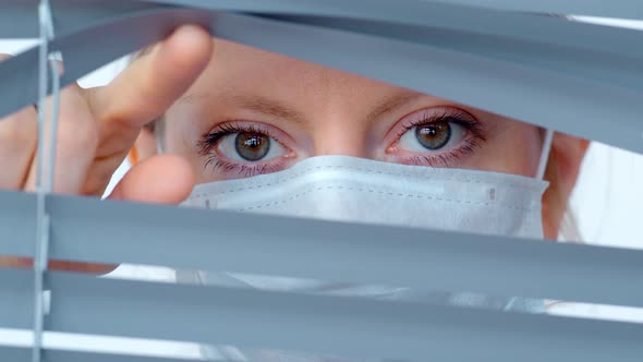 Young Woman in Medical Mask Looks Out the Window Through the Blinds