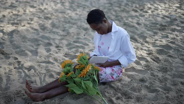 Young Relaxed African American Intelligent Woman Sitting on Sand with Bouquet of Flowers Reading