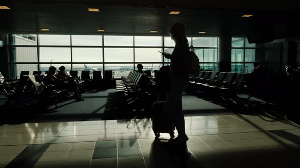 A Woman with a Bag on Wheels and a Phone in Her Hand Is Walking Along the Airport Terminal