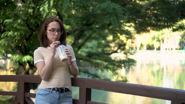 A Young Woman with Glasses Stands on a Bridge in the Park and Drinks Cold Coffee Closeup.