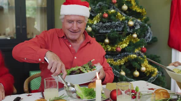 Happy caucasian senior man in santa hat serving himself food, at christmas dinner table with friends