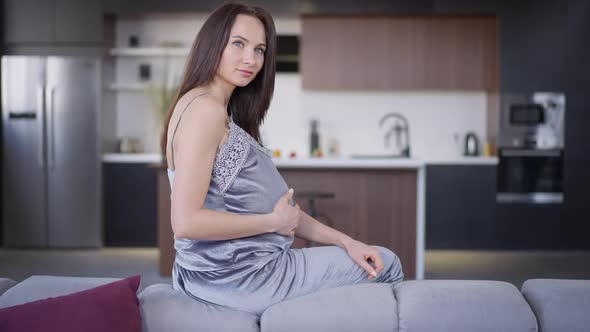 Confident Pregnant Beautiful Woman Sitting on Sofa Looking at Camera Caressing Belly