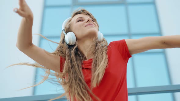 A Young Girl in Headphones is Listening to Music and Amusingly Dancing on a City Background