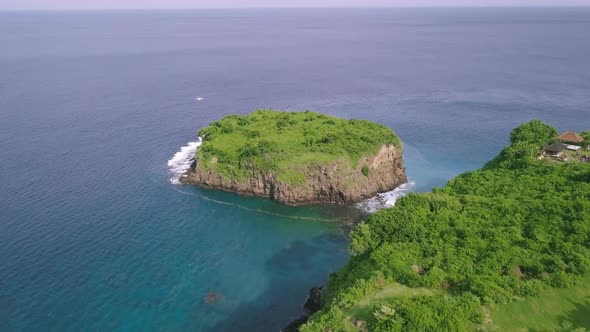 Drone Flight Over a Green Cliffside and a Green Islet at the Blue Sea.