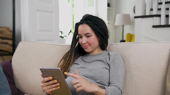Pregnant Woman with Dreadlocks Relaxing on the Soft Couch at Home and Browsing Photos on I-pad