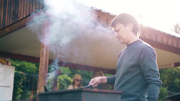 Young Man Standing Near the Summer House Prepares the Coals in the Grill The Girls Help Him