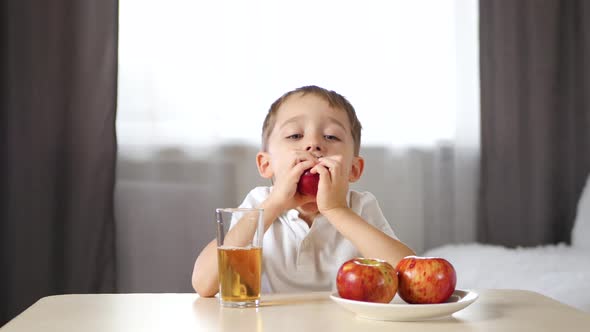 Happy Laughing Boy Sitting at the Table and Drinking Fruit Juice. The Child Bites Off a Slice of Red