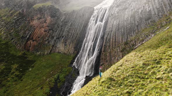 Happy Female Traveler Stands on a Picturesque Mountain Slope with Her Hands Raised Against the