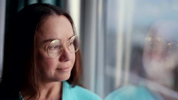 Pretty Middleaged Woman with Glasses Near Window is Looking at Camera and Smiling Kindly