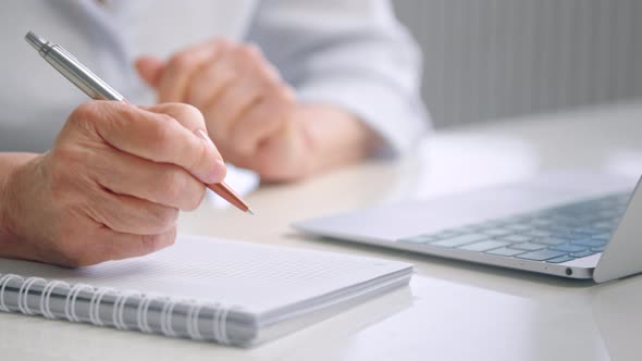 Senior lady teacher with wrinkly hands holds coloured pen over white page of paper notebook