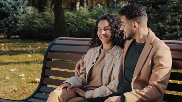 Cheerful Hispanic Couple Cutely Communicate Sitting on Bench in Autumn City Park Young Bearded Guy