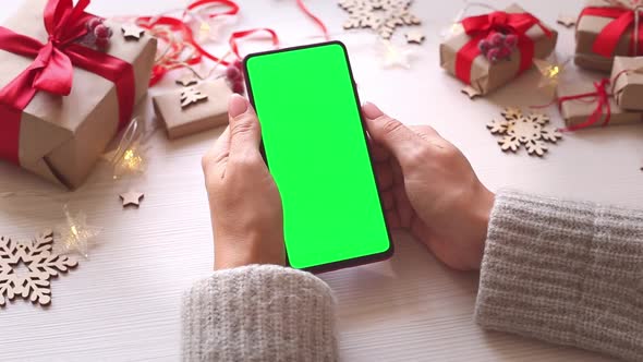 Woman holding smartphone with vertical green screen on Christmas background with gifts.