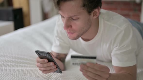 Young Man Using Credit Card on Smartphone in Bed