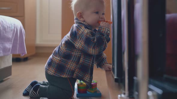 Small Boy Nibbles Bright Toy Siting on Floor at Rack in Room