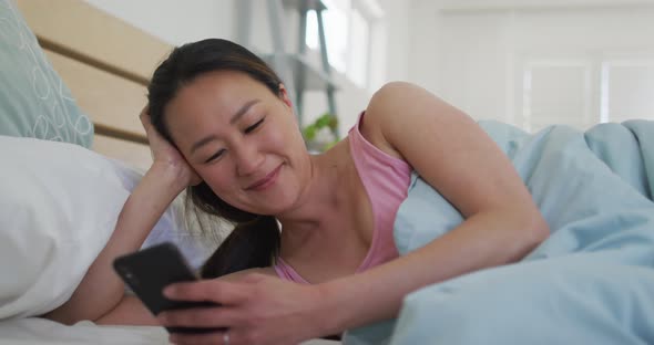 Happy asian woman lying in bed, using smartphone in the morning