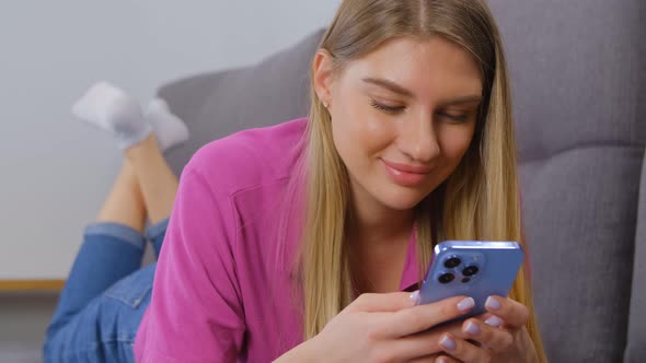 Young blonde woman typing message on modern smartphone while lying on couch