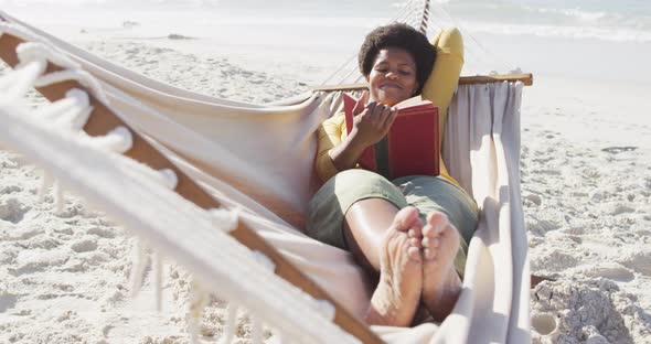 Happy african american woman reading and lying in hammock on sunny beach