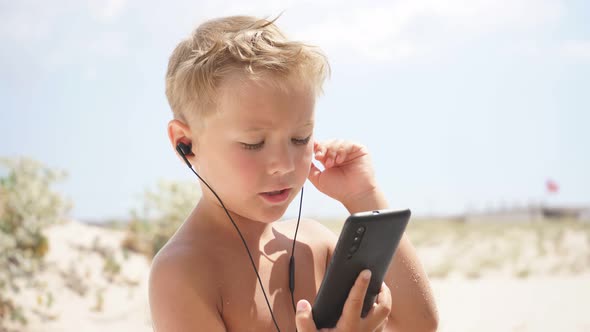 Tanned Boy in Headphones with a Smartphone in His Hands on the Beach