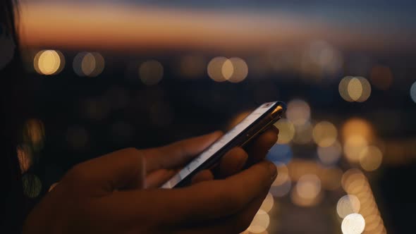 Close-up Female Hands Using Smartphone Messenger App at Stunning Bokeh Sunset City Lights Panorama