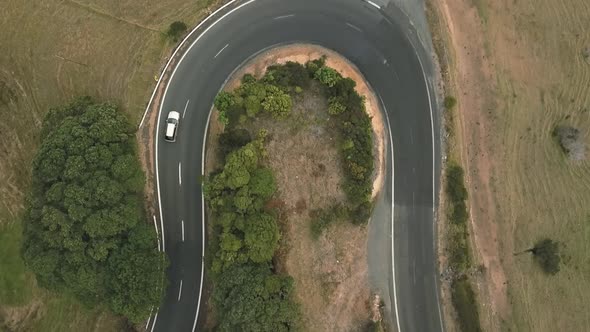 Aerial birds eye drone view of a white car driving round a bendy road in the hills of new zealand.