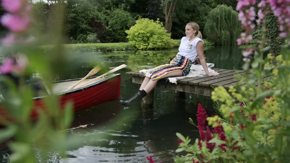 A Beautiful Woman Sitting On The Wooden Jetty By The Lake With A Boat Floating - full shot