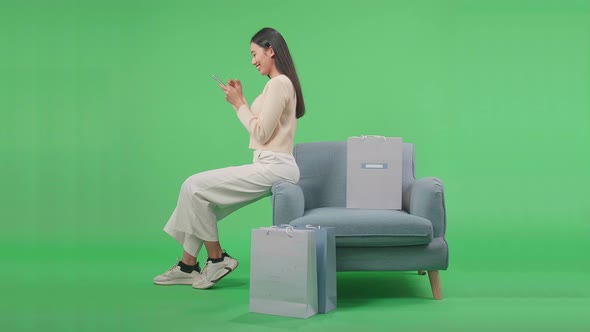 Woman With Shopping Bags Using Mobile Phone Shopping Online While Sitting On Sofa At Green Screen