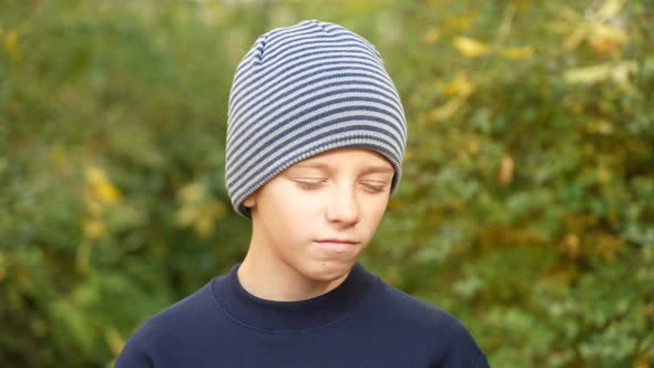 Serious Boy in Striped Hat Standing in Park Looks at Camera and Nods His Head in Disagreement
