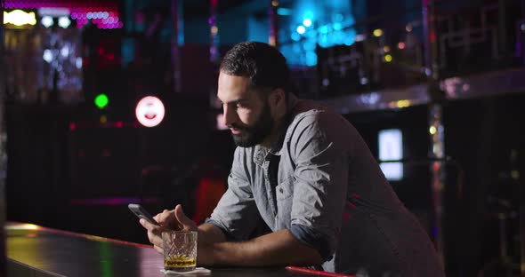 Young Middle Eastern Man Using Smartphone and Drinking Alcohol Next To Bar Counter in Night Club