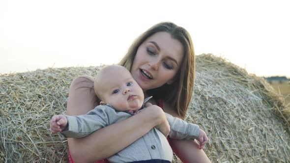 Young Mother Holds Her Baby Son When Leaned on Haycock in Spacious Field