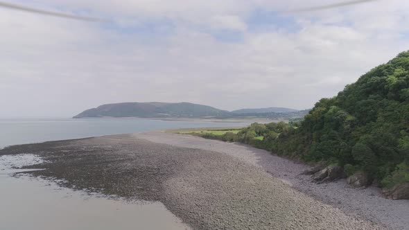 Wide aerial tracking forward over a pebbly beach, beside a prehistoric looking forest cliff, reveali