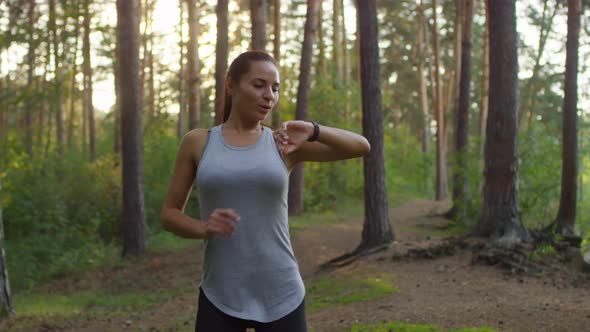 Cheerful Woman Running in Woods, Using Smartwatches and Posing for Camera